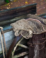 A collection of fishing nets draped over the edge of a wooden boat. The scene includes a mix of nets, ropes, and a rusty metal wheel, all set against a background of cobblestone paving with scattered leaves.