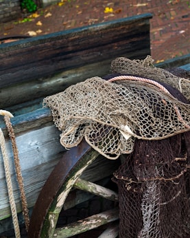A collection of fishing nets draped over the edge of a wooden boat. The scene includes a mix of nets, ropes, and a rusty metal wheel, all set against a background of cobblestone paving with scattered leaves.