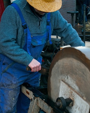 A person wearing blue overalls and a grey sweater operates a large circular grinding wheel. They are wearing a straw hat and appear to be focused on the task.
