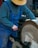 A person wearing blue overalls and a grey sweater operates a large circular grinding wheel. They are wearing a straw hat and appear to be focused on the task.