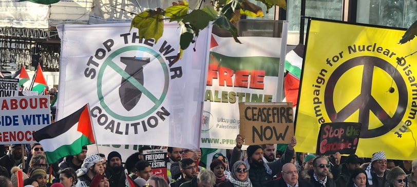 A group of protesters carrying various signs and banners advocating for peace and anti-war messages. Visible signs include calls for stopping wars, nuclear disarmament, and support for Palestine. People are holding Palestinian flags and banners with slogans like 'Stop the War Coalition', 'Ceasefire Now', and 'Free Palestine'. The gathering appears peaceful and crowded.