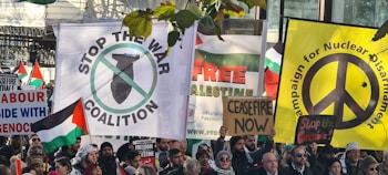 A group of protesters carrying various signs and banners advocating for peace and anti-war messages. Visible signs include calls for stopping wars, nuclear disarmament, and support for Palestine. People are holding Palestinian flags and banners with slogans like 'Stop the War Coalition', 'Ceasefire Now', and 'Free Palestine'. The gathering appears peaceful and crowded.