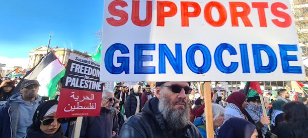 A group of people holding signs and banners during a protest. One sign reads 'SUPPORTS GENOCIDE' in red and blue, while another sign reads 'FREEDOM FOR PALESTINE' with additional text in Arabic. The crowd appears diverse, with multiple individuals wearing headscarves and winter clothing. Various flags, including Palestinian ones, are visible in the background.