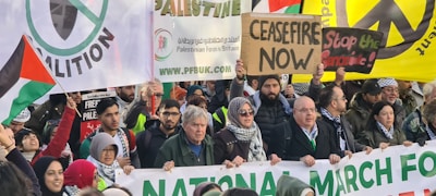 A diverse crowd of people participating in a protest, holding a variety of signs and banners advocating for ceasefire and peace. Many are wearing warm clothing and appear to be engaged in active discussion and demonstration. The image depicts a protest march, with signs in multiple languages and symbols indicating support for peace and Palestinian rights.