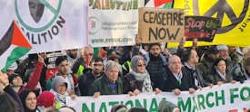 A diverse crowd of people participating in a protest, holding a variety of signs and banners advocating for ceasefire and peace. Many are wearing warm clothing and appear to be engaged in active discussion and demonstration. The image depicts a protest march, with signs in multiple languages and symbols indicating support for peace and Palestinian rights.