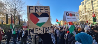 A group of protesters holding various signs and flags, with slogans like 'FREE PALESTINE' and 'Gaza Stop the massacre'. The scene takes place outdoors in what appears to be an urban setting, with buildings and trees visible in the background. The crowd is diverse, with many people wearing winter clothing and scarves.