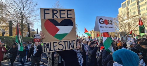 A group of protesters holding various signs and flags, with slogans like 'FREE PALESTINE' and 'Gaza Stop the massacre'. The scene takes place outdoors in what appears to be an urban setting, with buildings and trees visible in the background. The crowd is diverse, with many people wearing winter clothing and scarves.