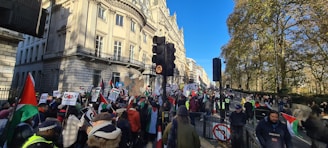 A large group of people gather in the street holding flags and signs, indicating a protest or demonstration. The backdrop features a historic building and trees, with a clear blue sky overhead. The crowd is diverse, and some police officers are present, ensuring safety and order.