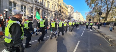 A group of police officers wearing high-visibility vests walk along a city street, with a demonstration taking place nearby. Flags and banners are visible among the crowd, which lines the sidewalk. The architecture of the buildings is historic, and trees line the street. The weather appears sunny, casting long shadows on the pavement.