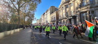 A group of people marching along a city street, some carrying flags and signs. Police officers in high-visibility jackets are present, overseeing the procession. The street is lined with large, historic buildings and trees. The scene appears to be captured in the daytime under clear skies.
