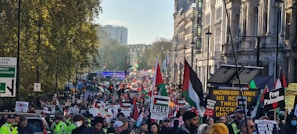 A large crowd of people gathered on a city street, holding signs and flags with messages of support for Palestine. The attendees are surrounded by urban buildings, trees, and signs indicative of a protest rally. Police officers are present to manage the crowd.