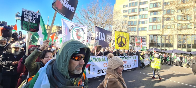 A large group of people are participating in a demonstration, holding various signs and banners. The banners express solidarity with Palestine, including messages for peace and justice. Many participants are bundled up in warm clothing, suggesting a cold day. The background includes a multi-story building and barren trees, indicating an urban setting.