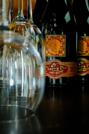 Several bottles of wine, specifically Cherry Pie Pinot Noir, are arranged with their labels visible. In front of the wine bottles, there are a number of empty wine glasses placed upside down on a dark surface. The background is slightly out of focus, emphasizing the bottles and glasses.