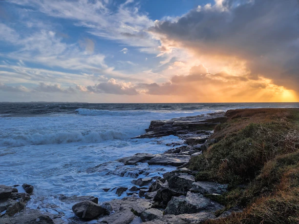 A rugged coastline at sunset with waves crashing against rocky shores under a moody sky.