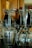A collection of upside-down wine glasses and beverage bottles, including a champagne bottle with a golden foil wrapping on top. The glasses and bottles are placed on a dark surface in front of a wooden paneled background, creating reflections and playing with light and shadows.