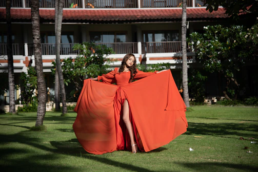 A graceful woman wearing a flowing ethnic dress, standing in a sunlit courtyard with lush greenery.