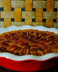 A rich pecan pie is displayed in a white dish with a red exterior. It is topped with neatly arranged pecan halves. The background is a textured, woven pattern composed of earthy tones, including shades of brown and yellow.