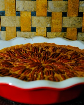 A rich pecan pie is displayed in a white dish with a red exterior. It is topped with neatly arranged pecan halves. The background is a textured, woven pattern composed of earthy tones, including shades of brown and yellow.