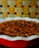 A rich pecan pie is displayed in a white dish with a red exterior. It is topped with neatly arranged pecan halves. The background is a textured, woven pattern composed of earthy tones, including shades of brown and yellow.