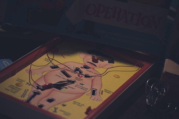 A vintage board game depicting a human body with labeled parts. The game involves removing pieces from the body using tweezers without touching the sides. It features a cartoonish character lying on an operating table, surrounded by various tools and labels like 'funny bone' and 'spare ribs'.