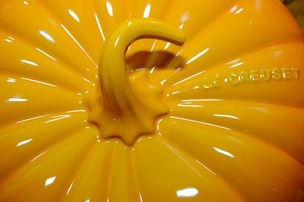 A close-up view of a bright orange glossy pumpkin-shaped object with the brand name 'Le Creuset' embossed on the surface. The image highlights the detailed ridges and the central handle, which resembles a pumpkin stem.