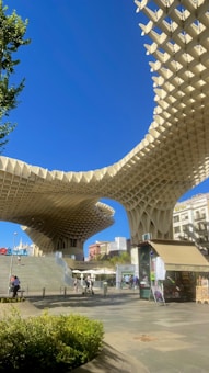 A modern architectural structure with a large, lattice-like wooden canopy stands prominently against a clear blue sky. The structure features curves and unique geometric patterns. Below, there are people walking and sitting on steps, enjoying the open space. Nearby, a kiosk or small shop with a canopy is visible, surrounded by greenery and other urban elements.