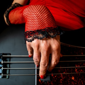 A hand with red nail polish playing a black guitar, adorned with a red fishnet glove edged with black lace. The background features a red garment, enhancing the dramatic contrast.