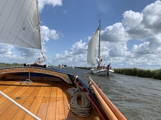 A serene scene featuring a tranquil sailing experience on a wide waterway. The foreground shows a boat with polished wooden flooring and decorative elements, while ahead, a sailboat with billowing white sails glides smoothly across the water. The sky is dotted with fluffy clouds, enhancing the peaceful atmosphere.