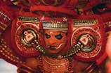 A vibrant and ornate costume featuring intricate patterns and bold colors, with a focus on a face adorned in traditional makeup and headgear. The detailed craftsmanship includes elaborate jewelry and symmetrical designs in red and gold tones.