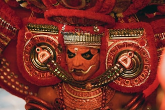 A vibrant and ornate costume featuring intricate patterns and bold colors, with a focus on a face adorned in traditional makeup and headgear. The detailed craftsmanship includes elaborate jewelry and symmetrical designs in red and gold tones.