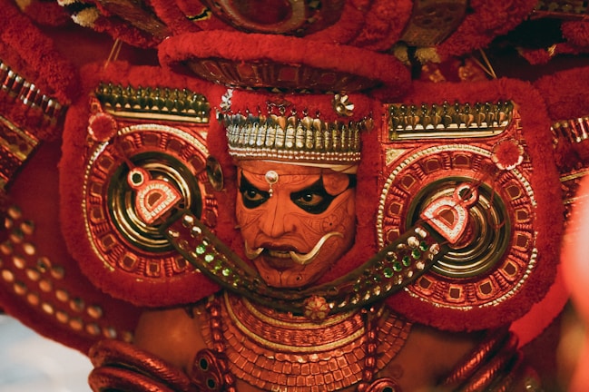 A vibrant and ornate costume featuring intricate patterns and bold colors, with a focus on a face adorned in traditional makeup and headgear. The detailed craftsmanship includes elaborate jewelry and symmetrical designs in red and gold tones.