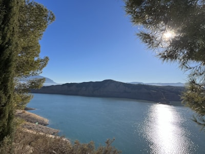 A vast, clear blue lake is surrounded by mountainous terrain. Sunlight glistens on the water's surface, creating a bright reflection. Tall trees frame the scene on both sides, adding a natural, serene atmosphere.