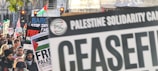 A protest featuring a crowd holding signs and banners related to Palestine solidarity and calls for a ceasefire. Several participants carry flags and placards with messages against military actions. Red traffic lights can be seen in the background, alongside trees and urban structures.