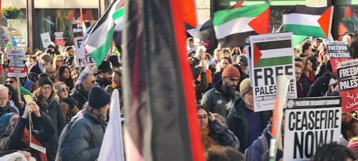 A large crowd of people is gathered, holding various signs and banners that express support for Palestine. Numerous flags, predominantly featuring red, white, green, and black colors, are being waved. The participants appear to be marching in a protest or rally, with signs displaying messages such as 'Free Palestine' and 'Ceasefire Now'. The crowd is diverse, with individuals dressed in winter clothing, suggesting cold weather.