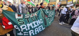 A group of people participating in a protest, holding large green banners with messages in white and red letters. One banner reads 'STOP ARMING ISRAEL' and another has 'SOLIDARITY WITH PALESTINE.' Some people are holding up various flags, and there are signs supporting Gaza. The protesters are walking on a city street bordered by buildings.