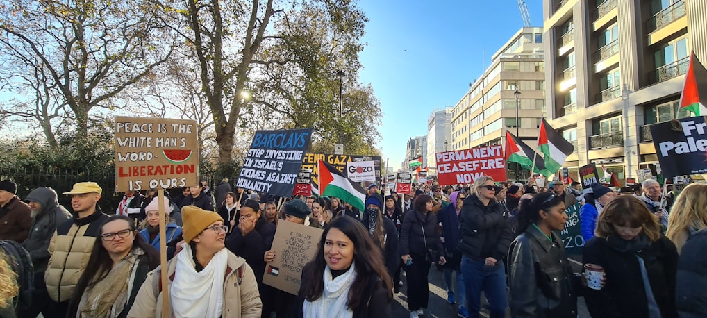 A large group of people are participating in a protest, holding various signs and flags. The signs carry messages advocating for peace, liberation, and a permanent ceasefire. Palestinian flags are visible among the crowd, which is diverse and bundled up in winter clothing. Trees and a modern building line the background on a sunny day.