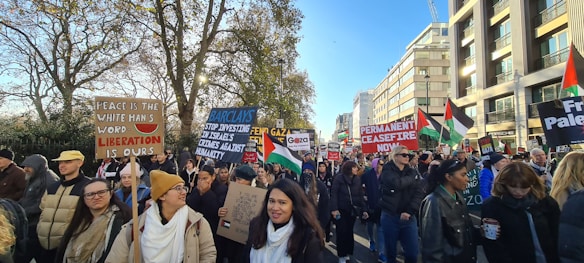 A large group of people are participating in a protest, holding various signs and flags. The signs carry messages advocating for peace, liberation, and a permanent ceasefire. Palestinian flags are visible among the crowd, which is diverse and bundled up in winter clothing. Trees and a modern building line the background on a sunny day.