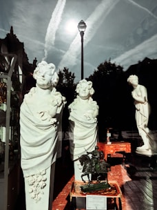 The image features statues and busts displayed in what appears to be a store window. There are three large marble busts of figures with intricate detailing and a full statue in the background. Reflections of a streetlamp and the sky with contrails are visible in the window glass. Additionally, there are some vintage items like a small statue of a knight on horseback and a decorative table also displayed.
