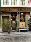 A quaint, vintage-style bookstore with a green façade features intricate wooden details and old-fashioned signage. The door is flanked by tall windows, and a wooden bench outside bears the words 'This is my happy place.' A potted plant is positioned beside the entrance, adding a touch of nature to the scene.