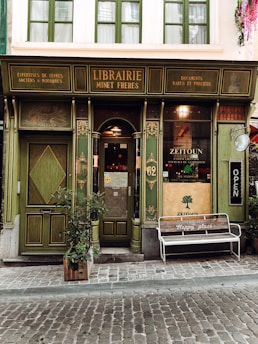 A quaint, vintage-style bookstore with a green façade features intricate wooden details and old-fashioned signage. The door is flanked by tall windows, and a wooden bench outside bears the words 'This is my happy place.' A potted plant is positioned beside the entrance, adding a touch of nature to the scene.