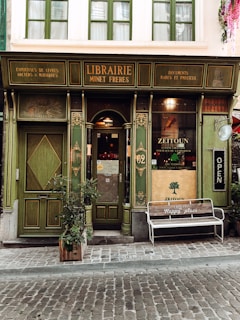 A quaint, vintage-style bookstore with a green fa&ccedil;ade features intricate wooden details and old-fashioned signage. The door is flanked by tall windows, and a wooden bench outside bears the words 'This is my happy place.' A potted plant is positioned beside the entrance, adding a touch of nature to the scene.