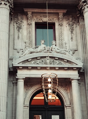 An ornate architectural facade featuring classical elements such as fluted columns and elaborate stone carvings. Two stone statues depicting reclining figures adorn the area above an arched doorway, which is topped with intricate relief work. A modern light fixture, composed of a vertical series of concentric rings containing several light bulbs, hangs prominently in front of the structure.