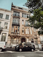 A multi-story residential building with ornate architectural details and large windows. There are two vehicles parked on the street in front of the building. One van is white, and the other is black. A leafy tree partially shades the view of the building.