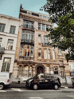 A multi-story residential building with ornate architectural details and large windows. There are two vehicles parked on the street in front of the building. One van is white, and the other is black. A leafy tree partially shades the view of the building.