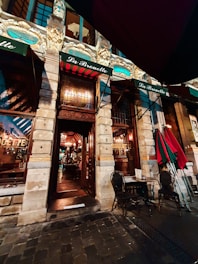 The image depicts an ornate storefront of a restaurant or café, featuring detailed architectural elements and decorative moldings. There are two green and red striped awnings with the name 'La Brouette' written on them. The entrance is flanked by stone columns, with two chairs and a small round table set up outside on the cobblestone pavement.