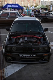 A black BMW car with its hood open, showcasing the engine. The car is parked on a tiled pavement at an outdoor car show or gathering, with other vehicles and a crowd in the background. Tents and banners are set up around the event space.