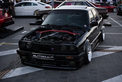 A sleek black car is prominently displayed in an urban setting with its hood open, revealing a modified engine with red accents. The vehicle sits on a stylized pavement marked with white geometric patterns. In the background, other cars and a crowd of people can be seen, indicating a car event or show.
