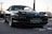 A sleek, dark-colored BMW sports car with pop-up headlights is parked in an outdoor setting. The car's license plate reads 'AUTOCULT,' and it is positioned on a paved area. The background reveals a gathering of vehicles and people, possibly at a car show or meetup.