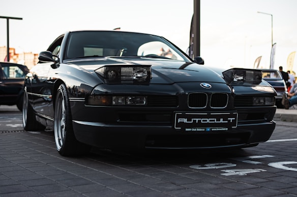 A sleek, dark-colored BMW sports car with pop-up headlights is parked in an outdoor setting. The car's license plate reads 'AUTOCULT,' and it is positioned on a paved area. The background reveals a gathering of vehicles and people, possibly at a car show or meetup.