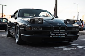 A sleek, dark-colored BMW sports car with pop-up headlights is parked in an outdoor setting. The car's license plate reads 'AUTOCULT,' and it is positioned on a paved area. The background reveals a gathering of vehicles and people, possibly at a car show or meetup.